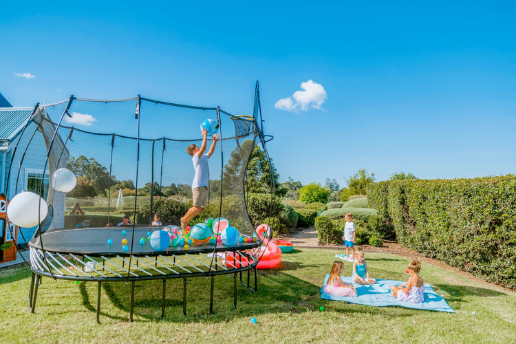 Children playing on a trampoline in a backyard with a clear blue sky.
